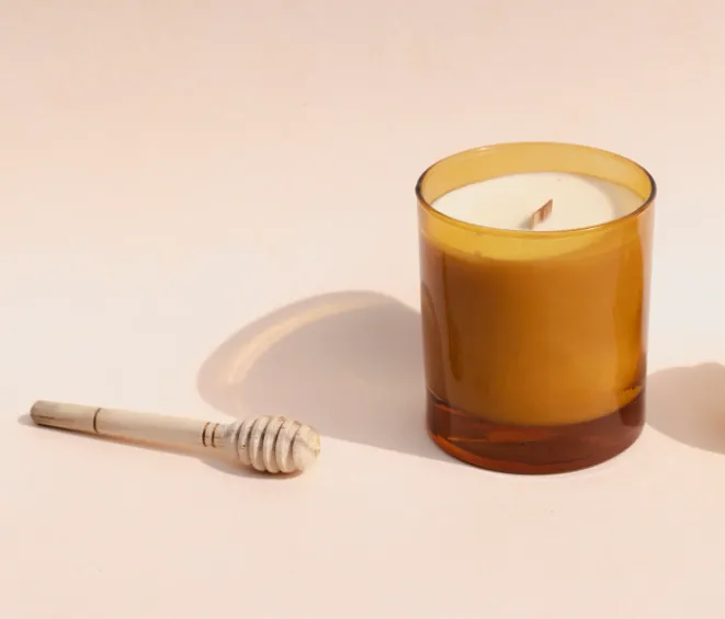 Candela in cera d'api in un vasetto ambrato, accanto a un mestolino da miele